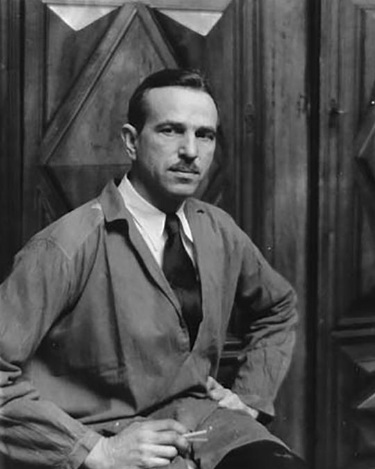 Black and White photo of a man wearing a overcoat with a tie. He has a widow's peak and a mustache. 