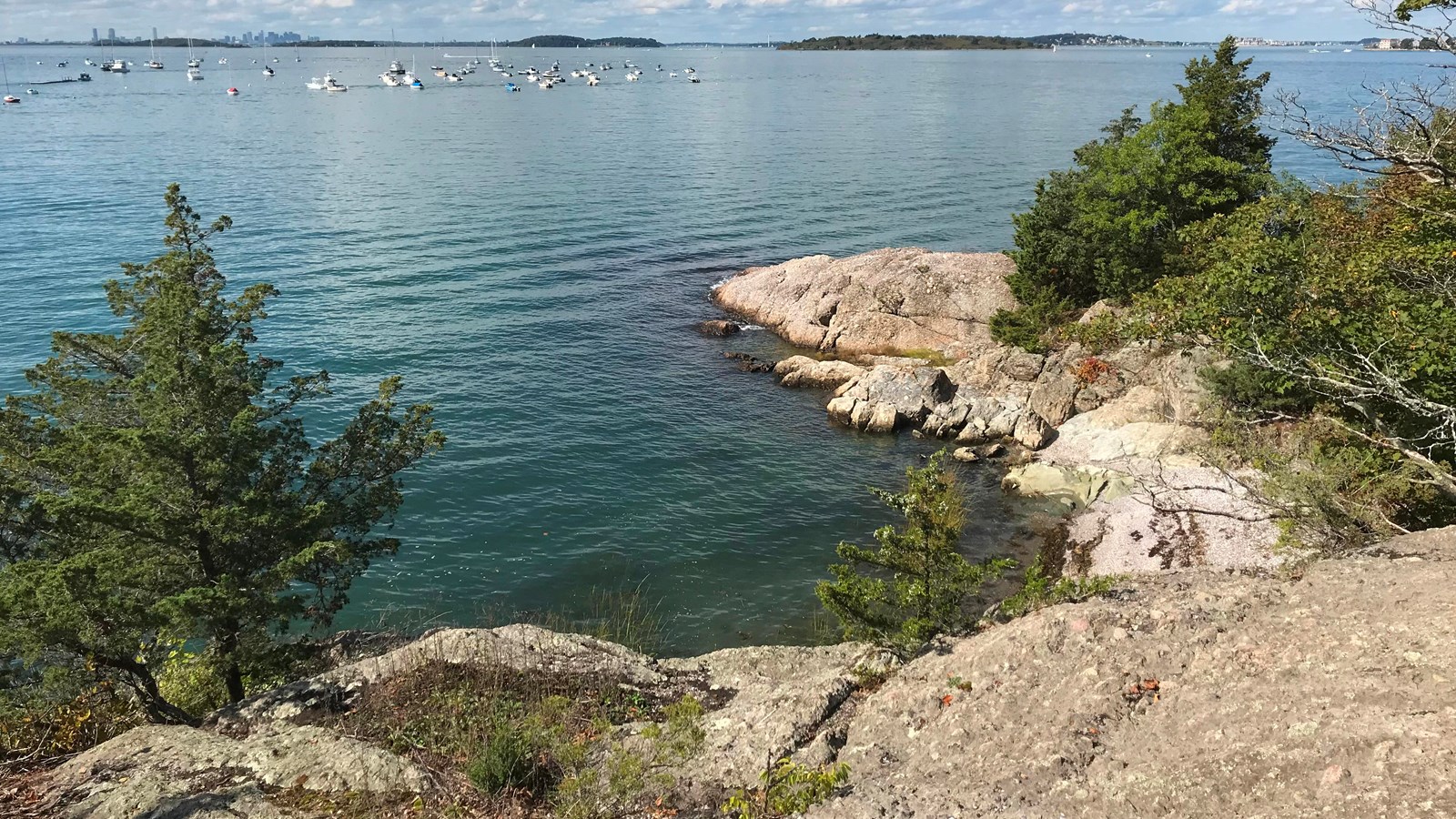 A rocky coast in the foreground lining teal green harbor with boats in the distance. 