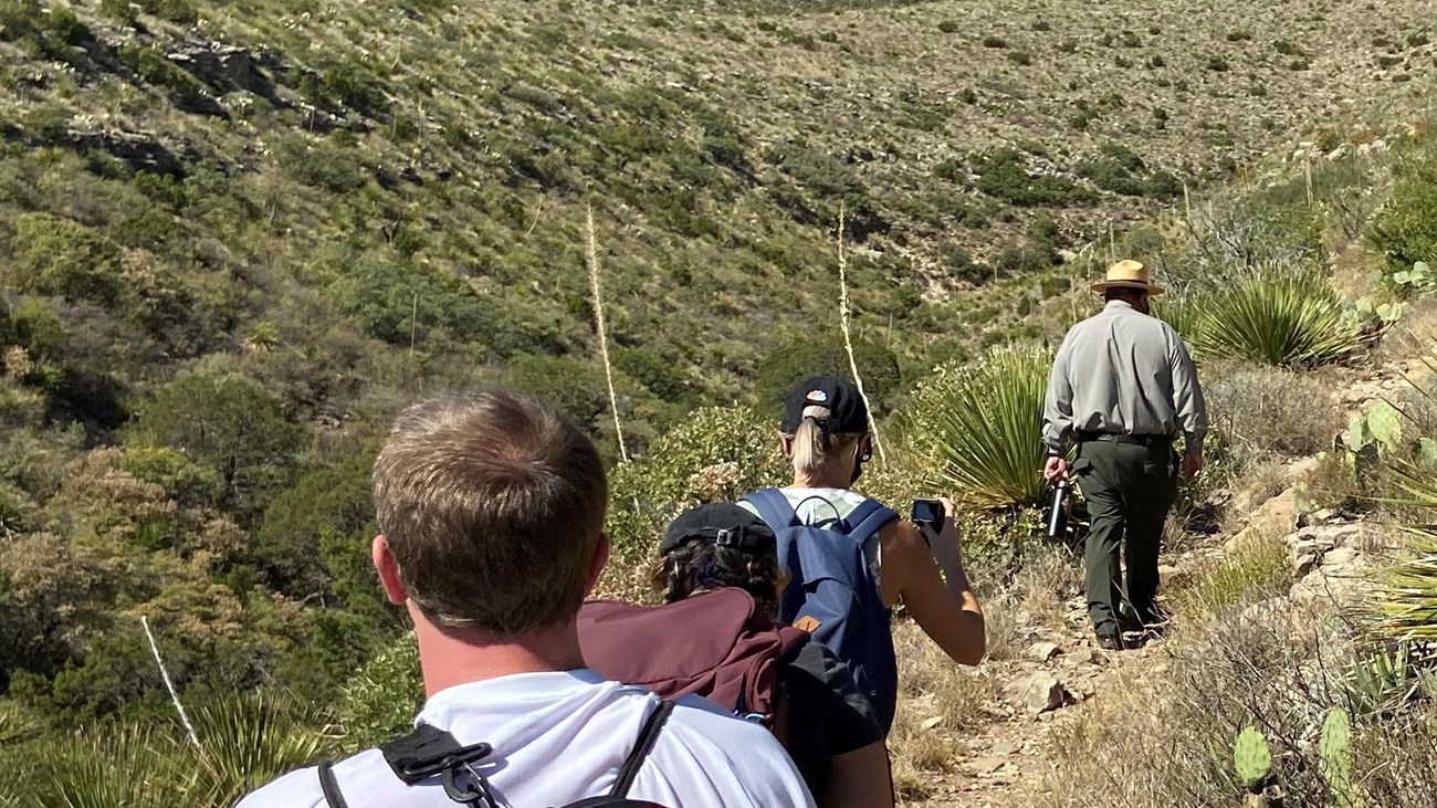 A uniformed park ranger and visitors hike up a desert trail