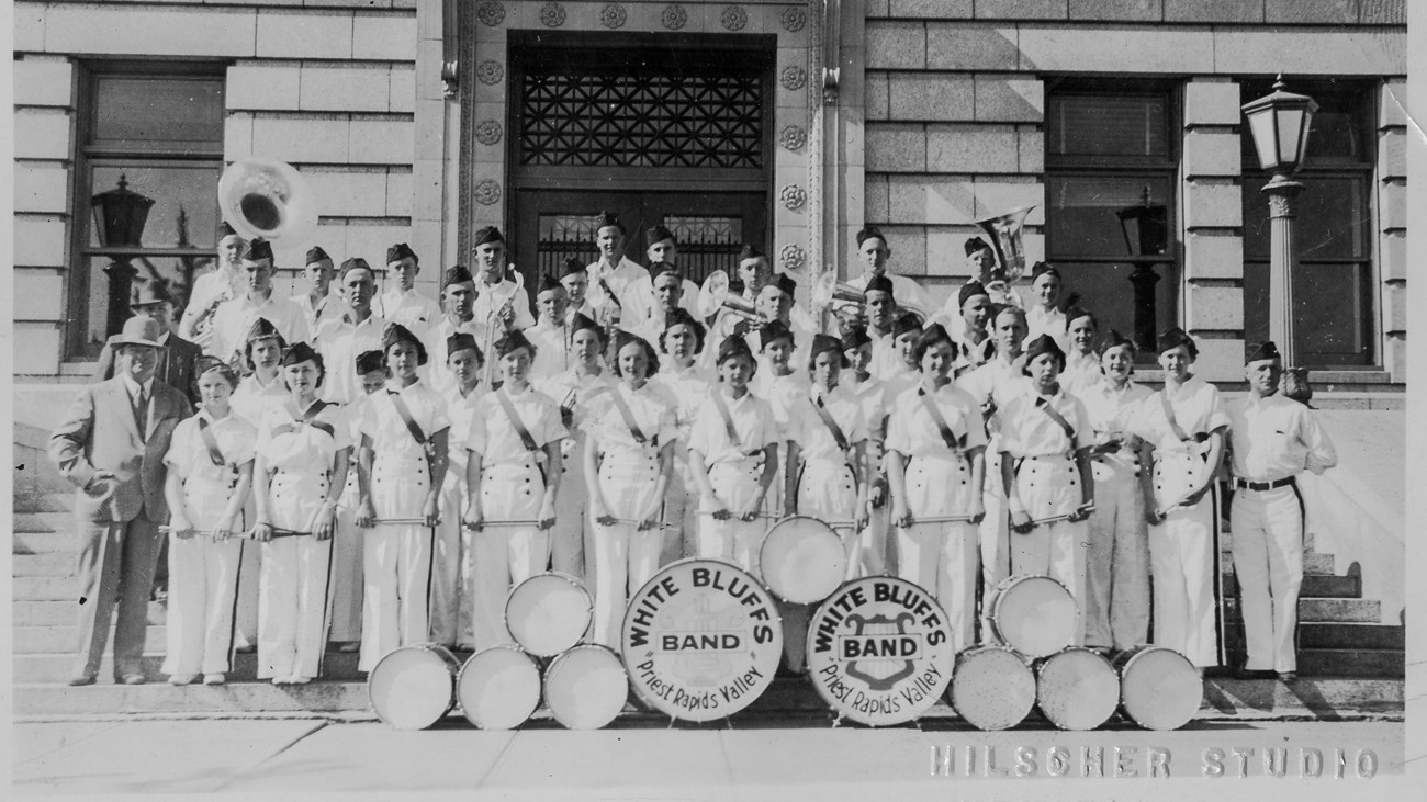 Historic photo of a school band in uniforms standing in front of a  brick building