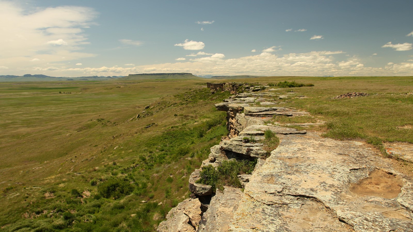 A sandstone cliff descends into open grasslands with mountains in the distance
