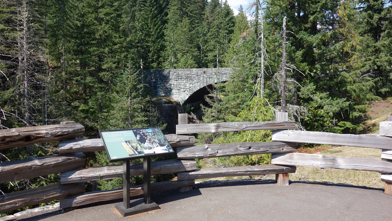 A wayside in a metal frame next to split-rail fence overlooking a canyon with a stone bridge.