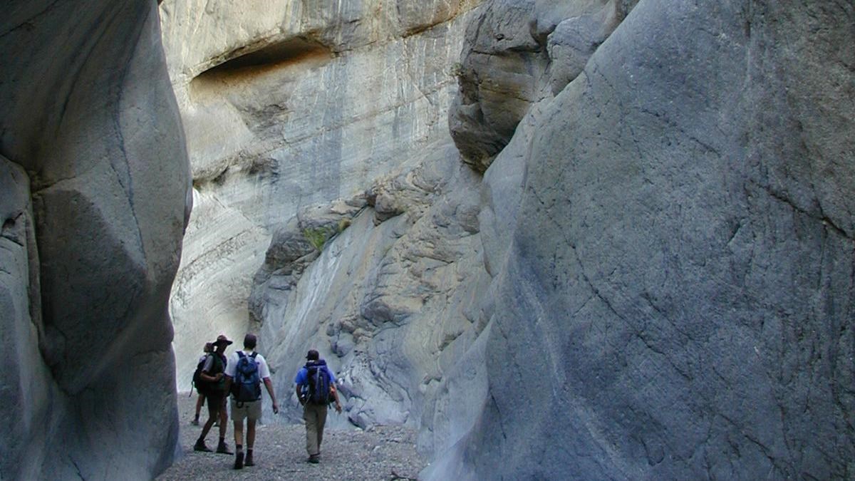 Hikers are dwarfed by smooth, near-vertical walls in a shaded canyon.