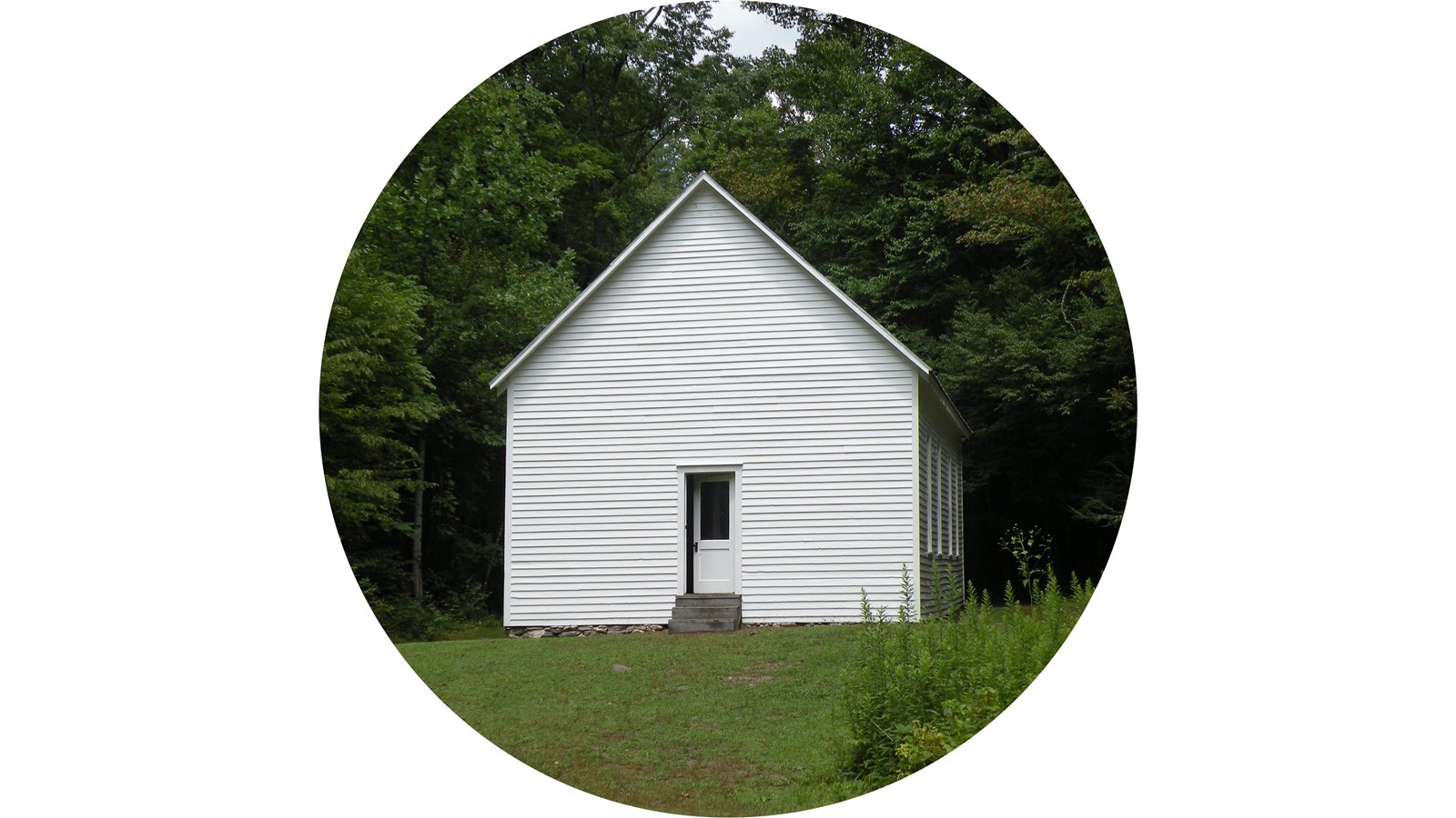 A white schoolhouse with a grassy lawn surrounded by dark green trees.