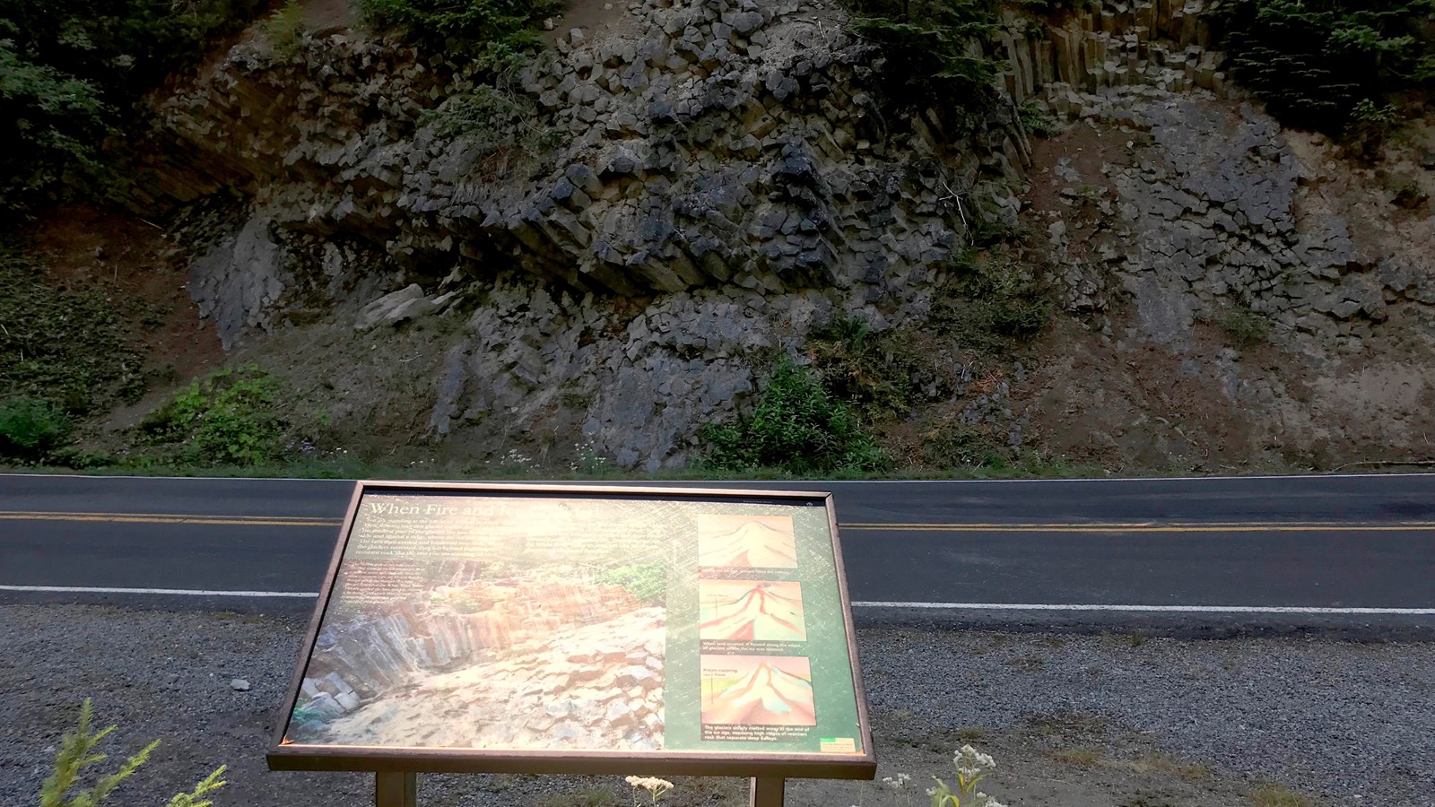 A wayside panel overlooks a road towards a rock formation exposed in a hillside.