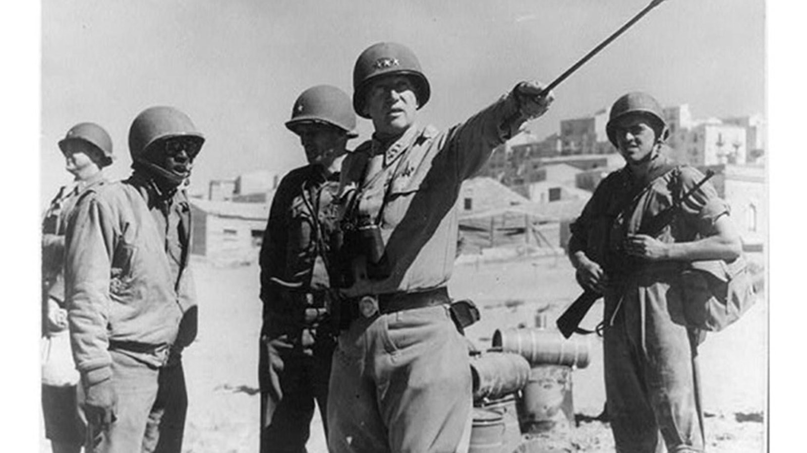 Man in uniform standing with sunglasses on. He is surrounded by other uniformed men.