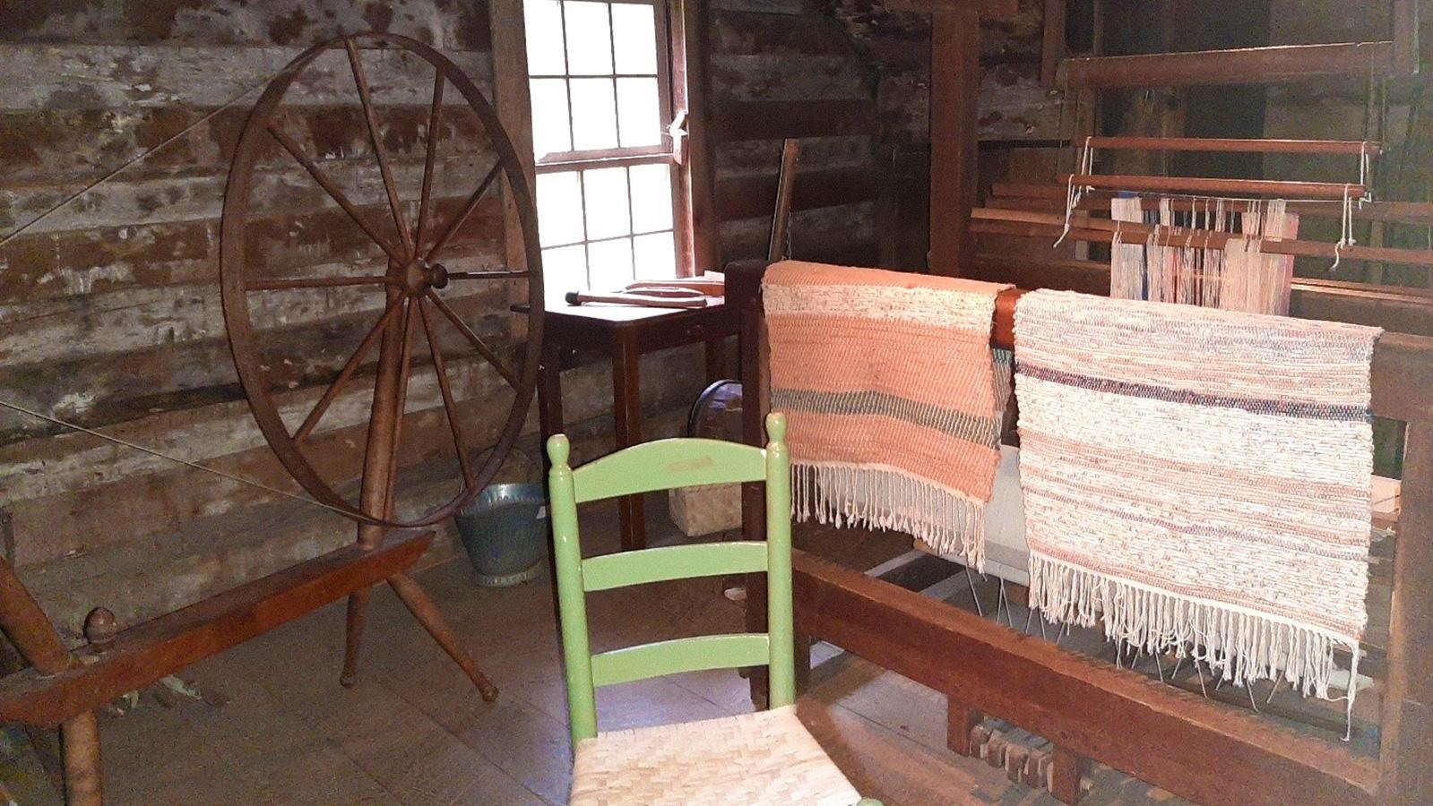 A wooden chair and spinning wheel sit beside 2 woven rugs slung over a rack inside a rough cabin