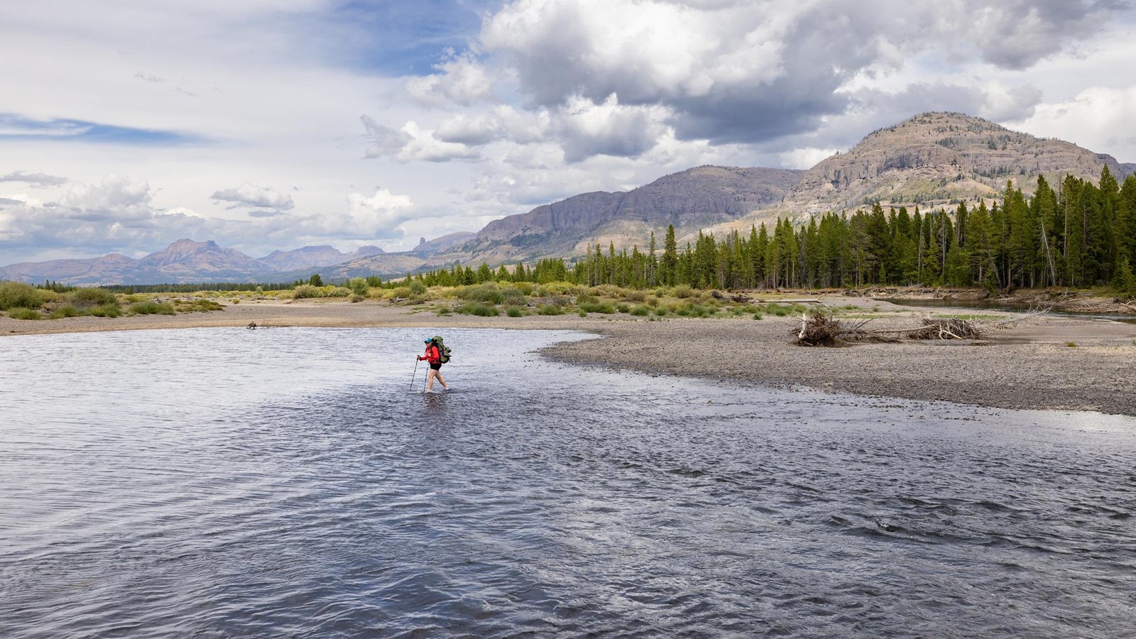 A hiker fords a river with mountains in the background.