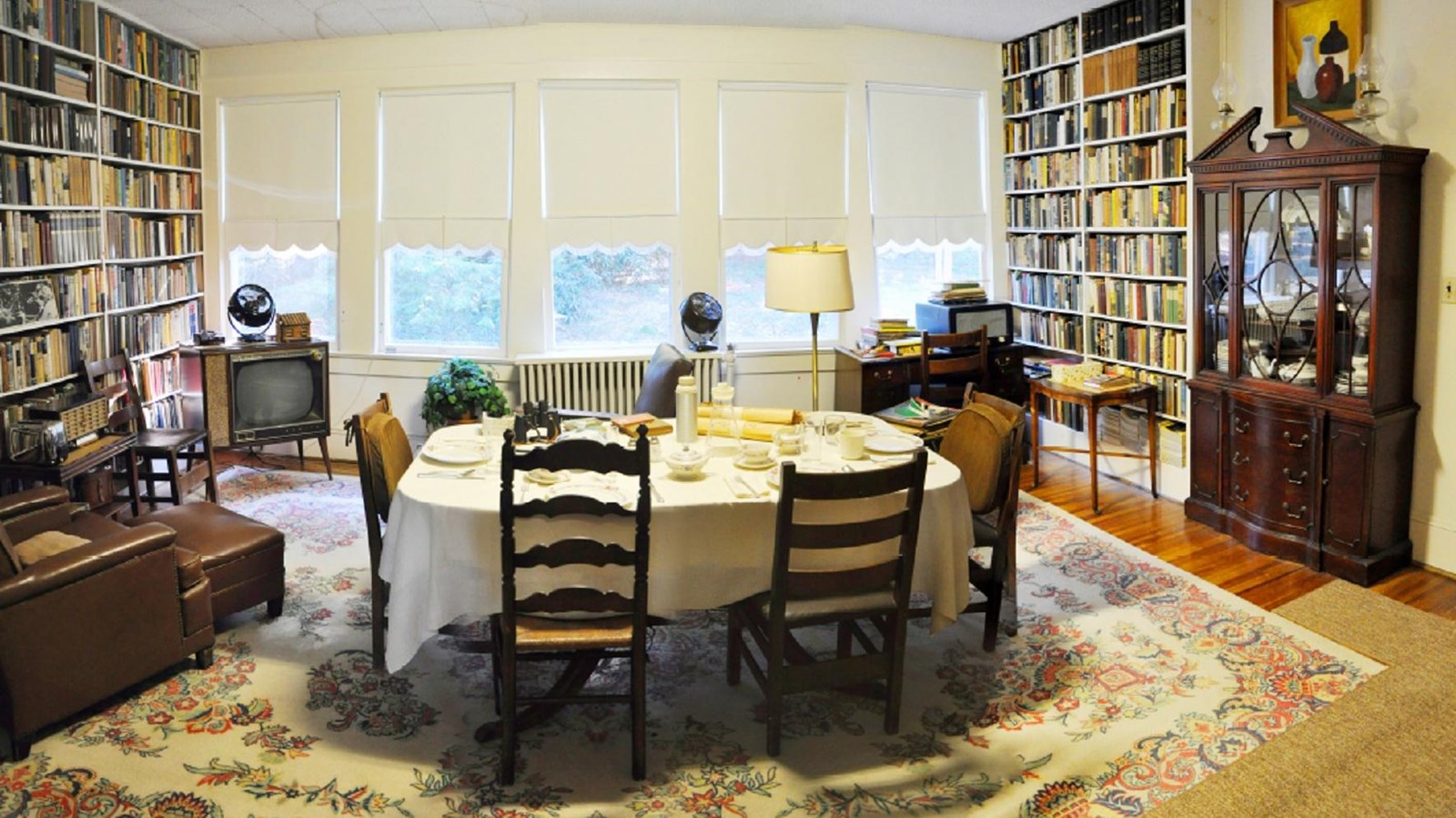 Windows, a dining table with four mix matched chairs and a TV sits between two filled bookcases, a