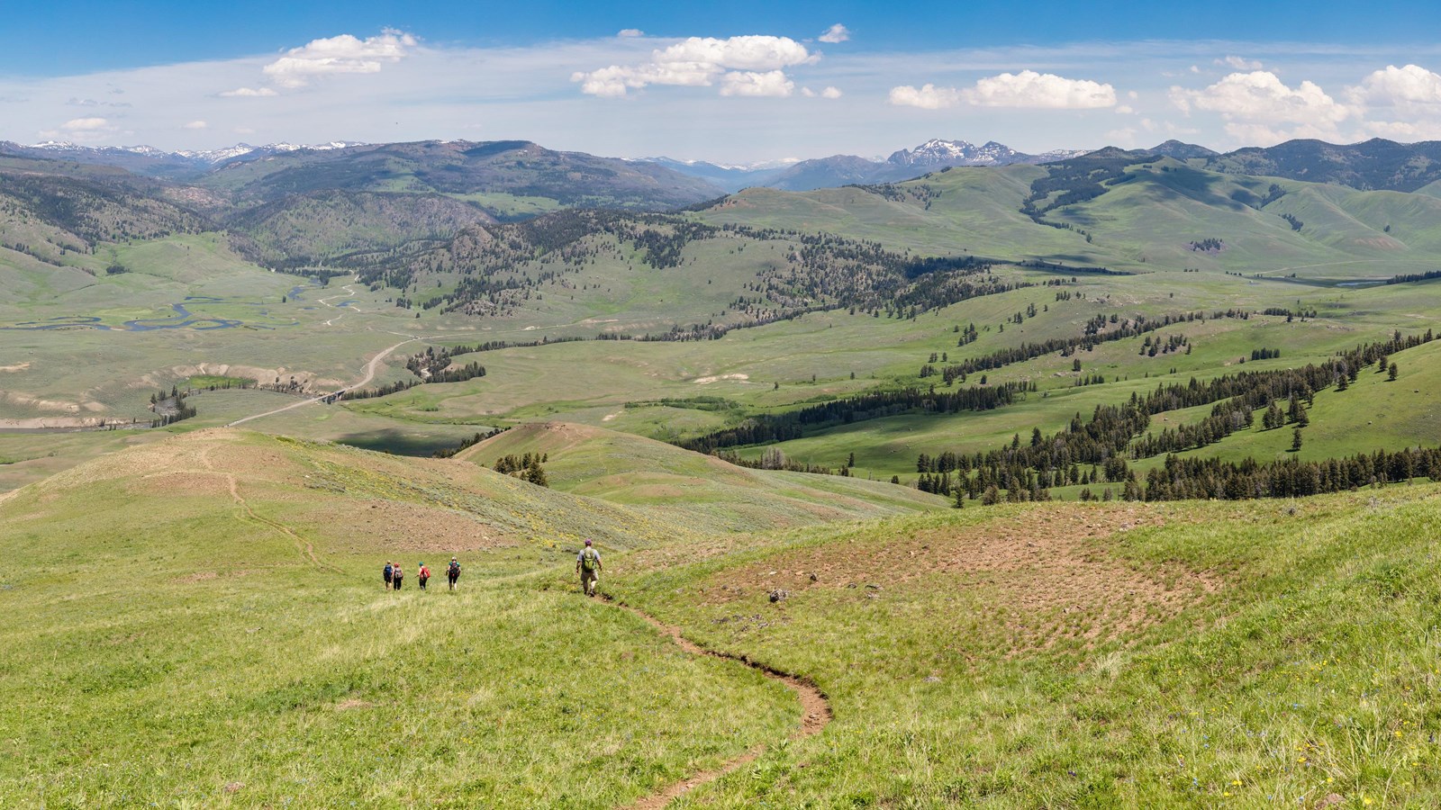 Hikers travel down a trail along a green slope to a valley below.