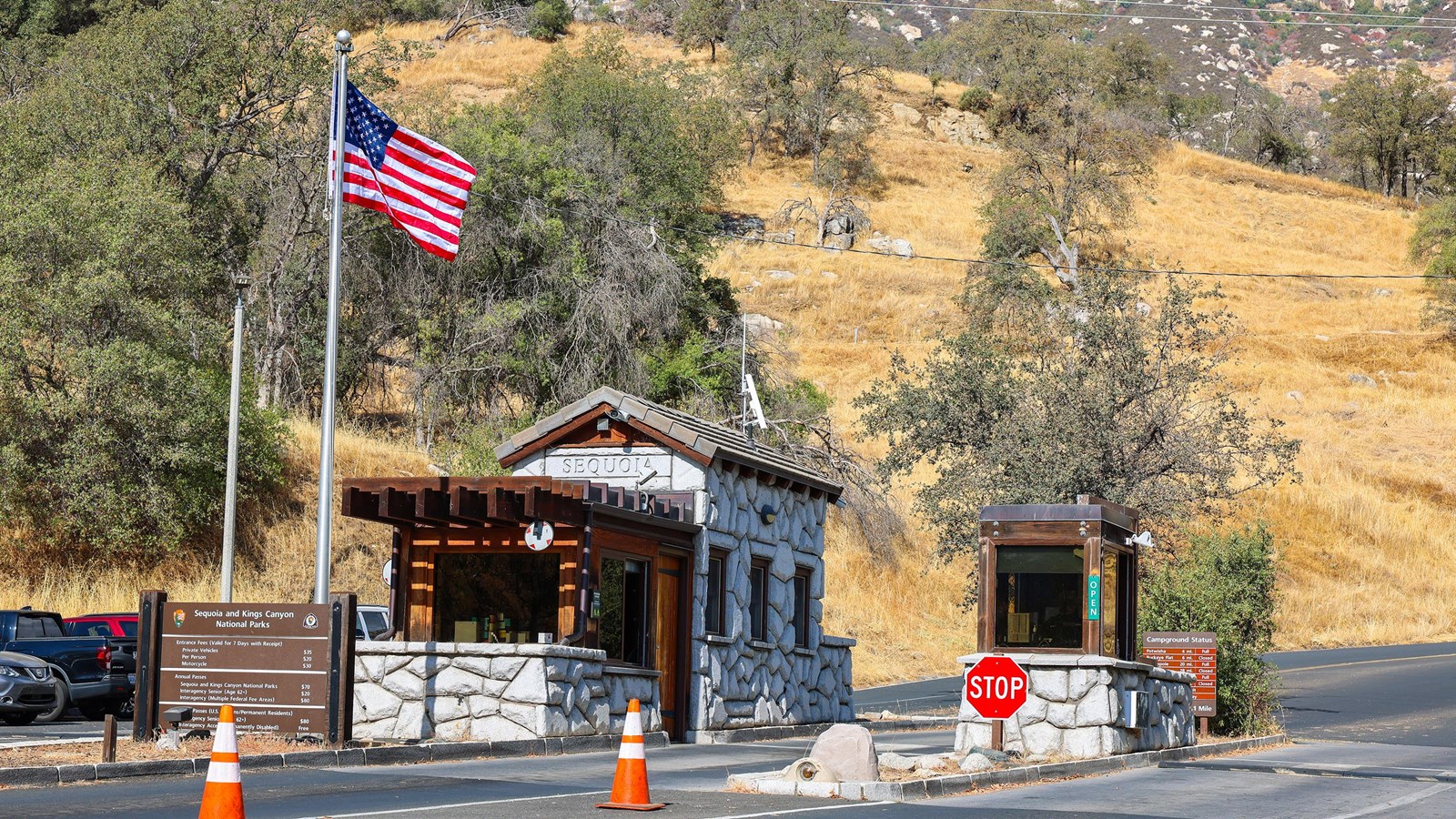 Two small rock and wood building with windows next to a flagpole in the middle of a roadway