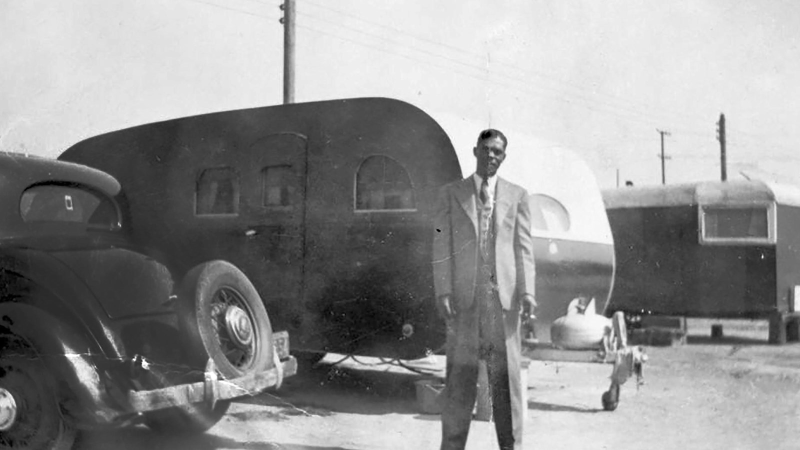 A historic photo of an African American man in a suit standing in front of a mobile trailer home. 
