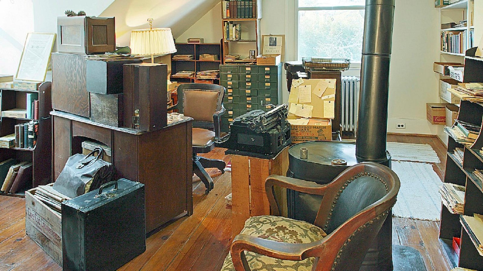A small desk in the center, is surrounded by loaded bookshelves, stacks of paper, an old wood stove.