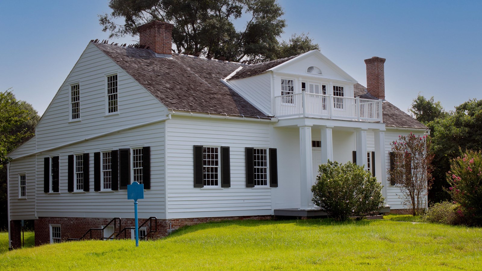 A white house with black shudders and cedar shake roof behind green grass.