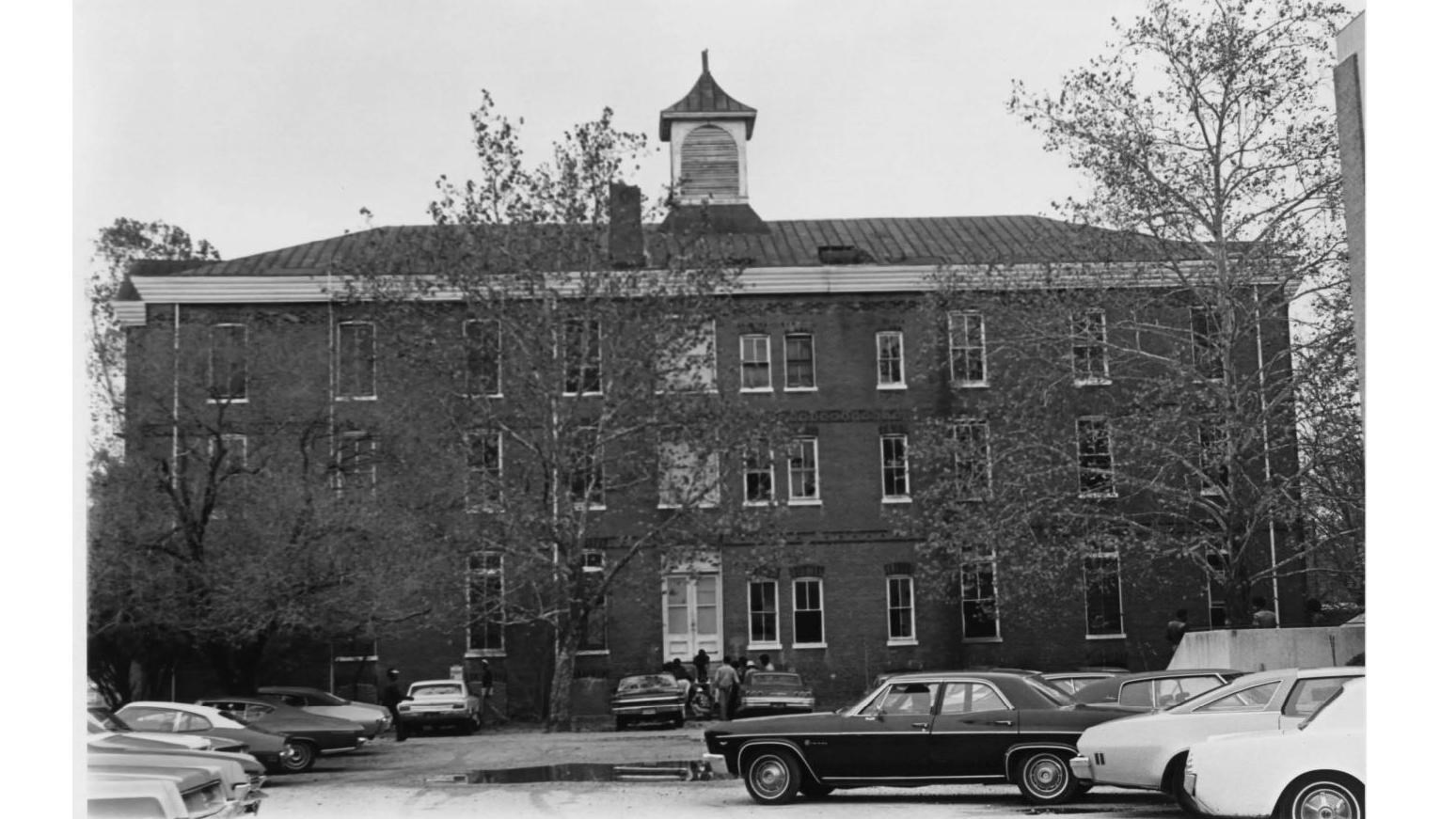 tall symmetrical academic building with a parking lot full of cars