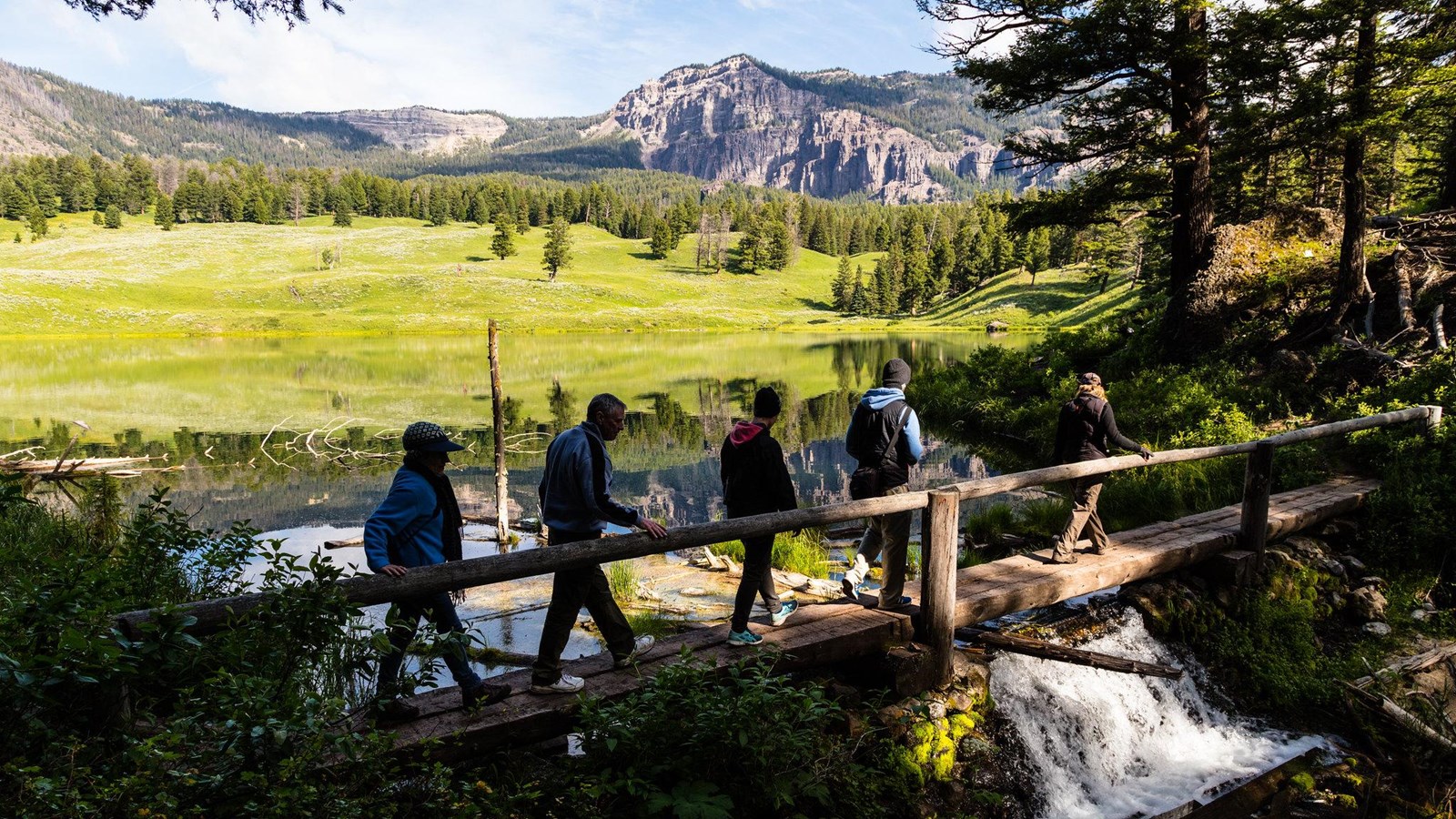 Hikers walk across a bridge at the outlet of a small lake surrounded by rolling hills.
