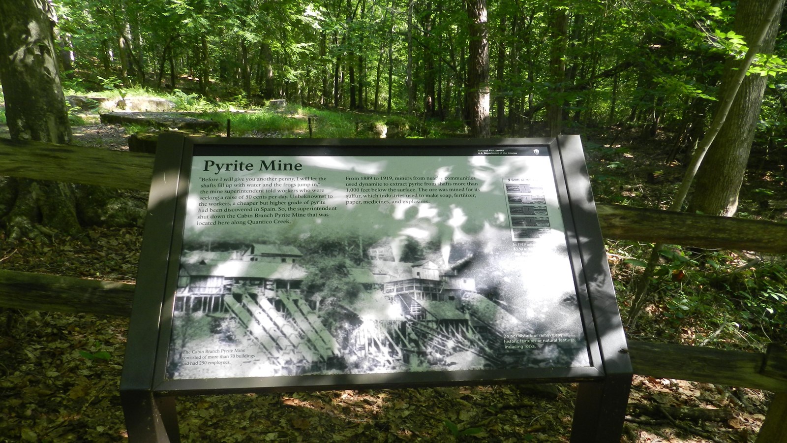 Sign at trail edge in front of fence protecting low squares of concrete foundations in the forest
