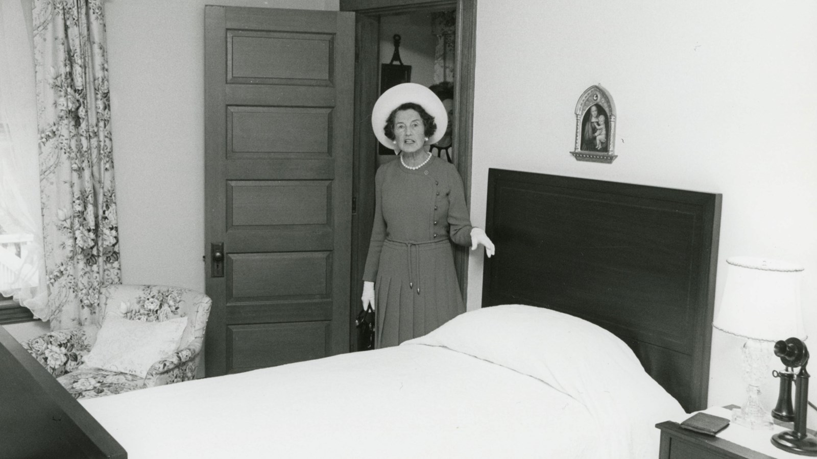 Woman in large white hat stands next to twin bed