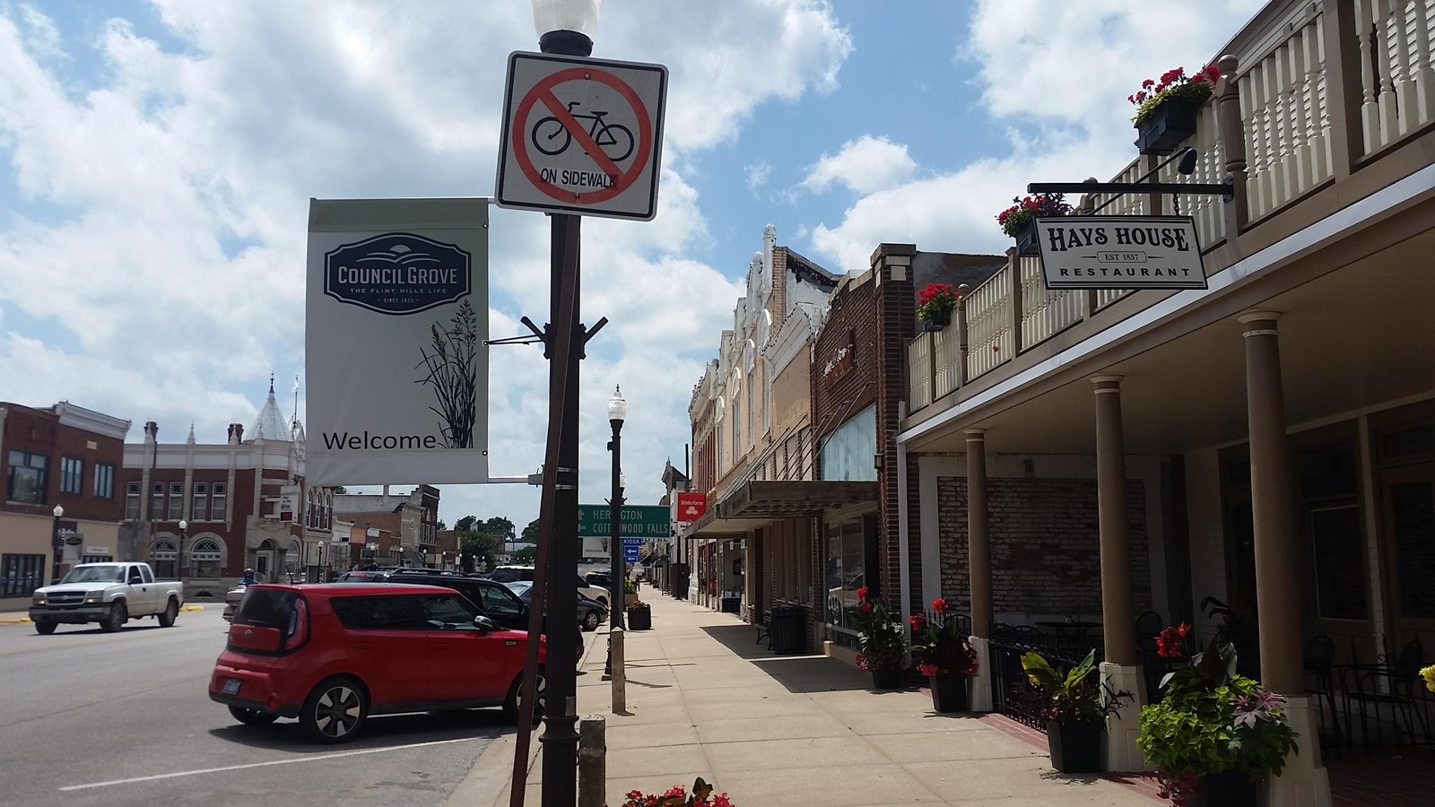 View down a small-town main street with buildings with potted plants out front, a sidewalk, cars, an