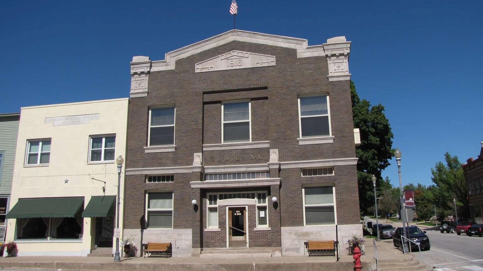 A two story brick bank building with a flag on the roof occupies a downtown corner.