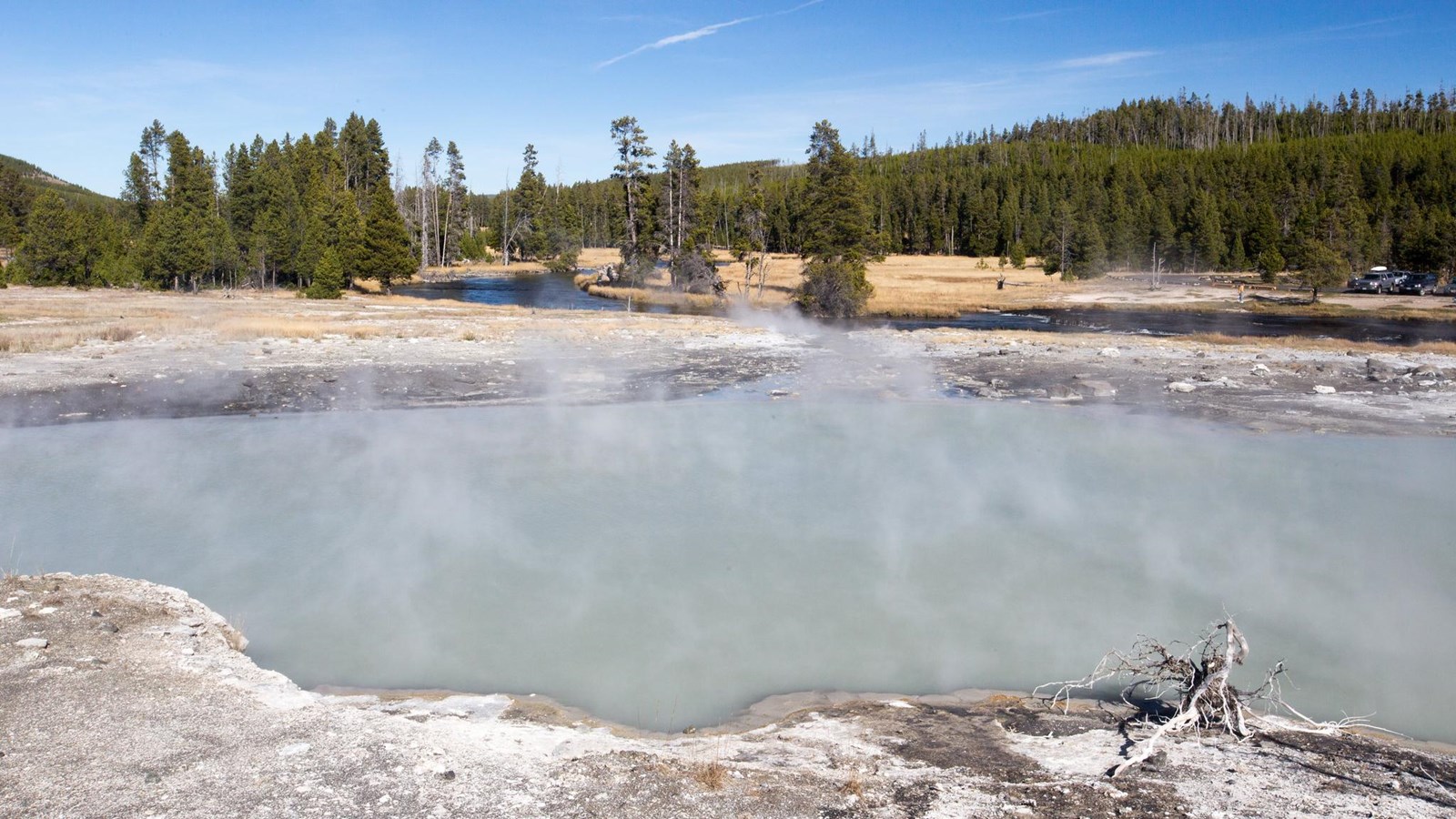 Steam rises off of a large, gray hot spring located near a river.