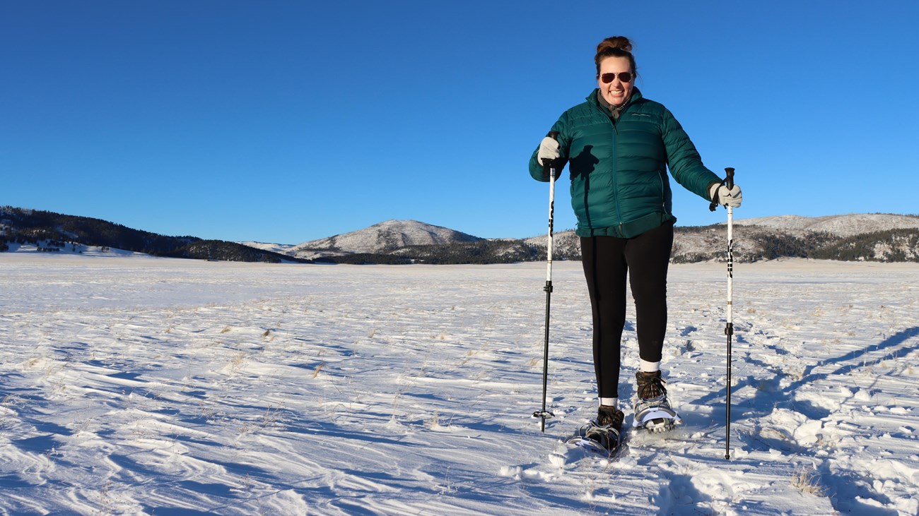 A woman in a green coat smiles as she snowshoes toward us.