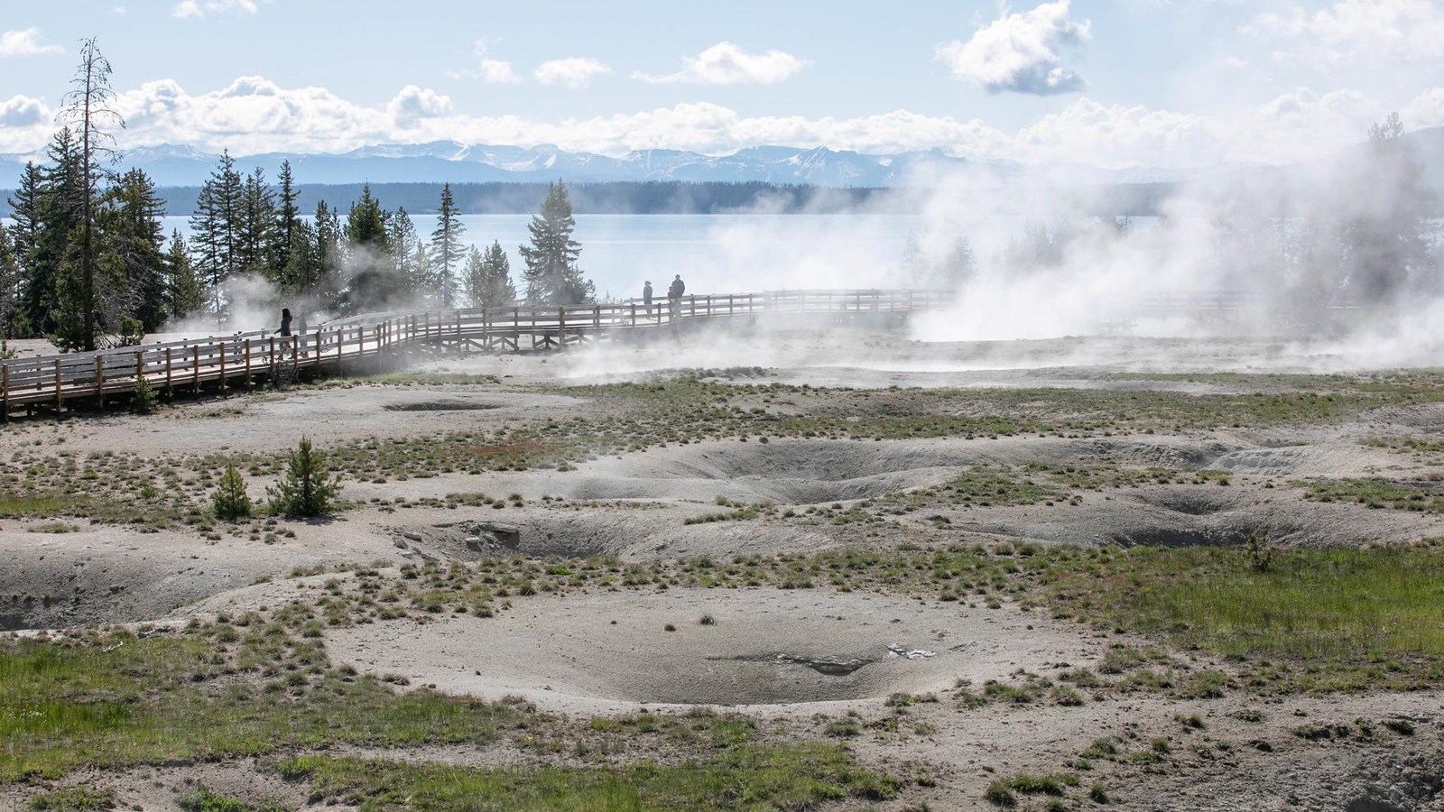 Steam rises off of thermal features in a geyser basin on the shore of a large lake.