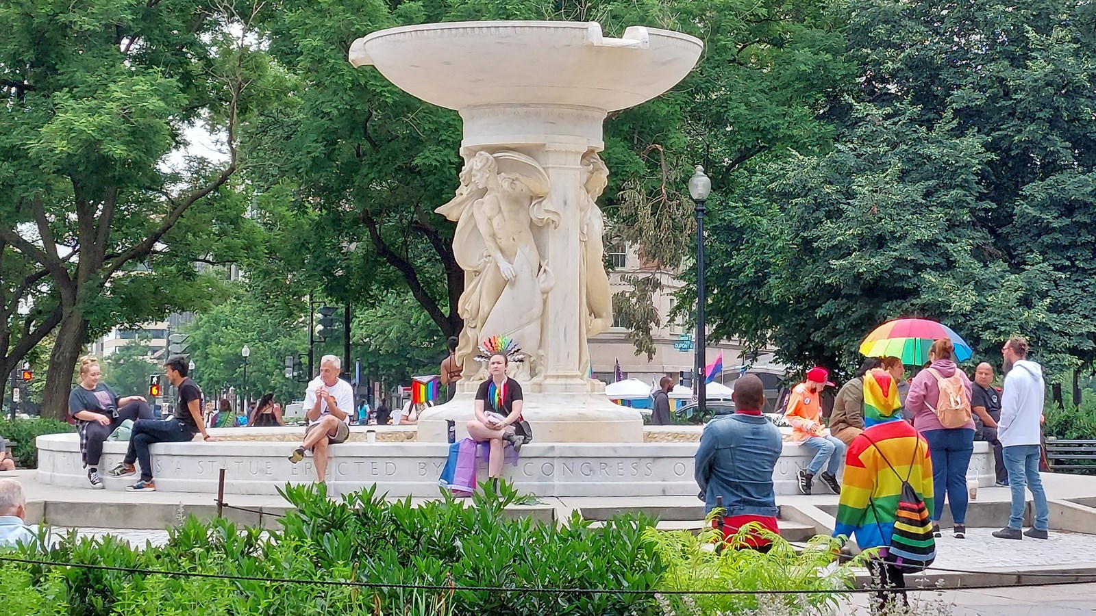 Fifteen people, some wearing rainbow accessories sitting and standing at the Dupont Circle fountain