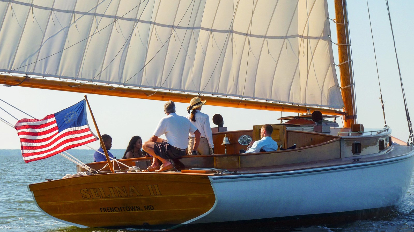 A sailboat with a large white sail on a calm body of water near a marina, under a partly cloudy sky.