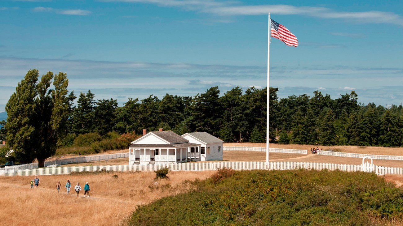 Historic white buildings in a grassy field. Hikers are going along various trails.