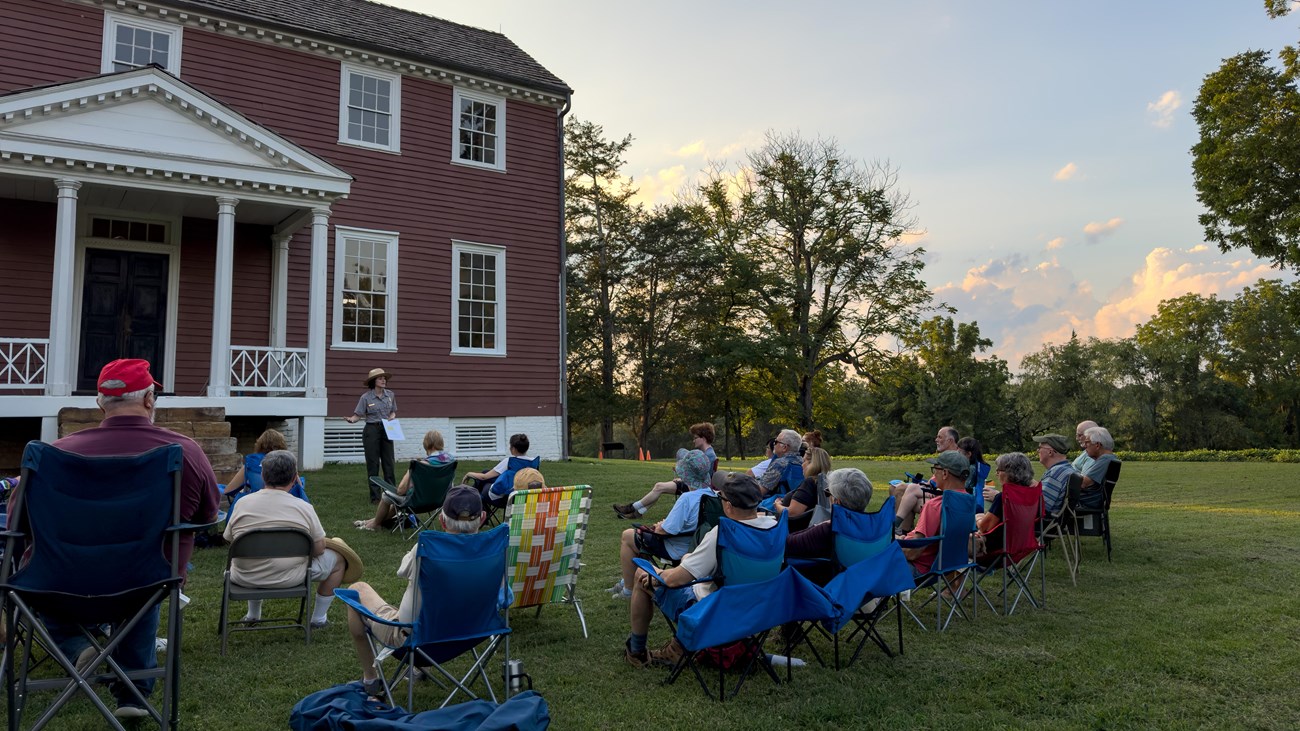 A ranger speaks to a group of 30 people seated on a lawn in front of a historic house.