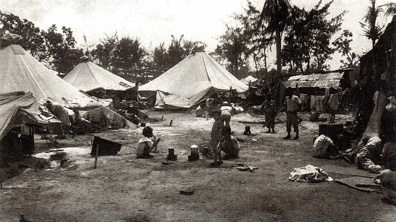 3 children squat near tin cans. 3 tents are to the left. Laundry hangs from lines attached to tents.