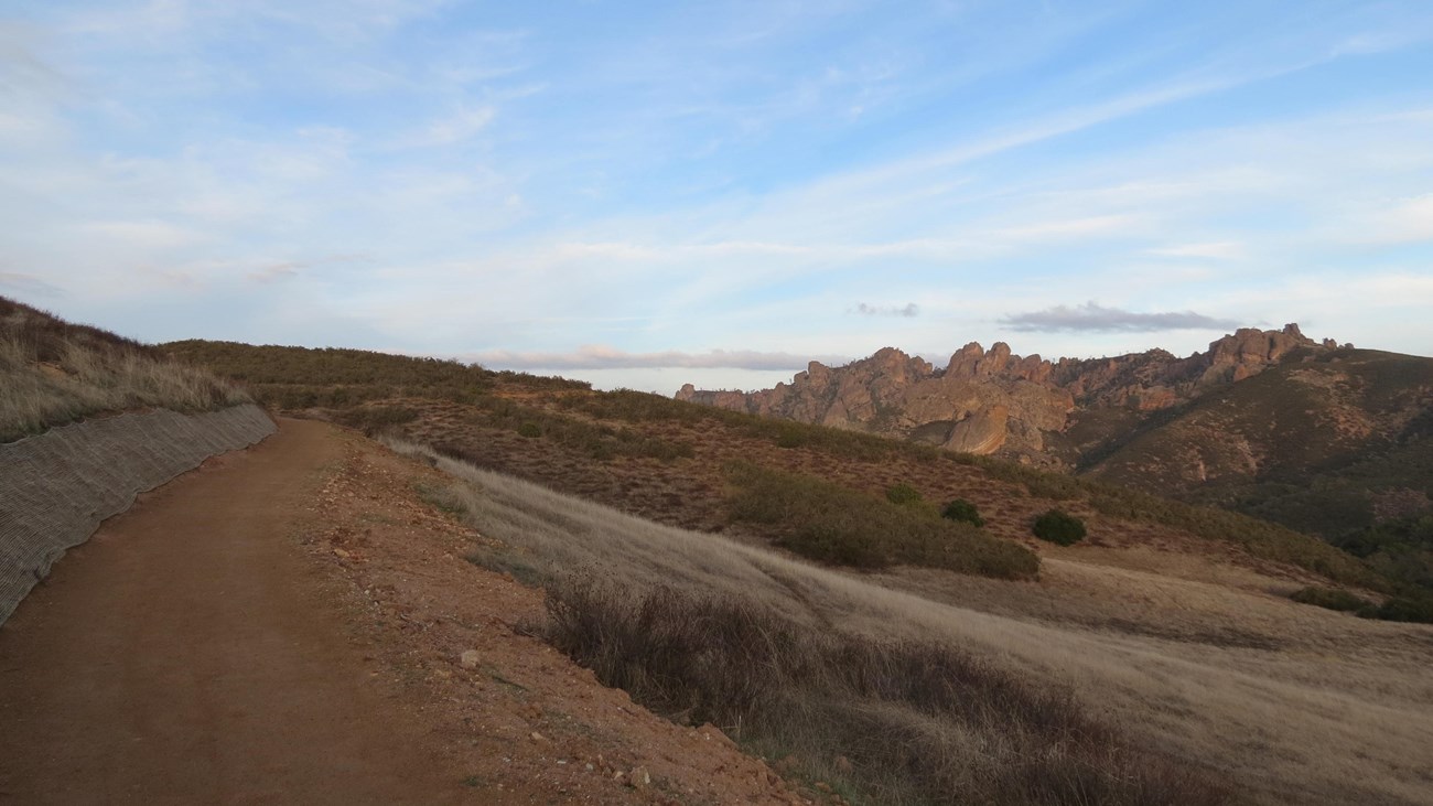 Hard pack dirt trail winding around hillside, large rock spires appearing in the distance.