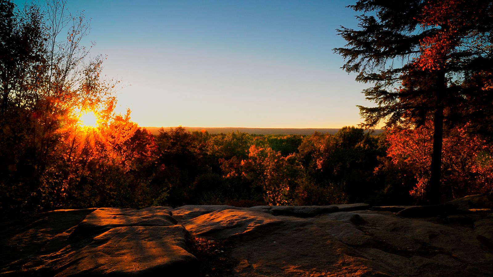 View from a rocky ledge looking out at a sunset over fall treetops.