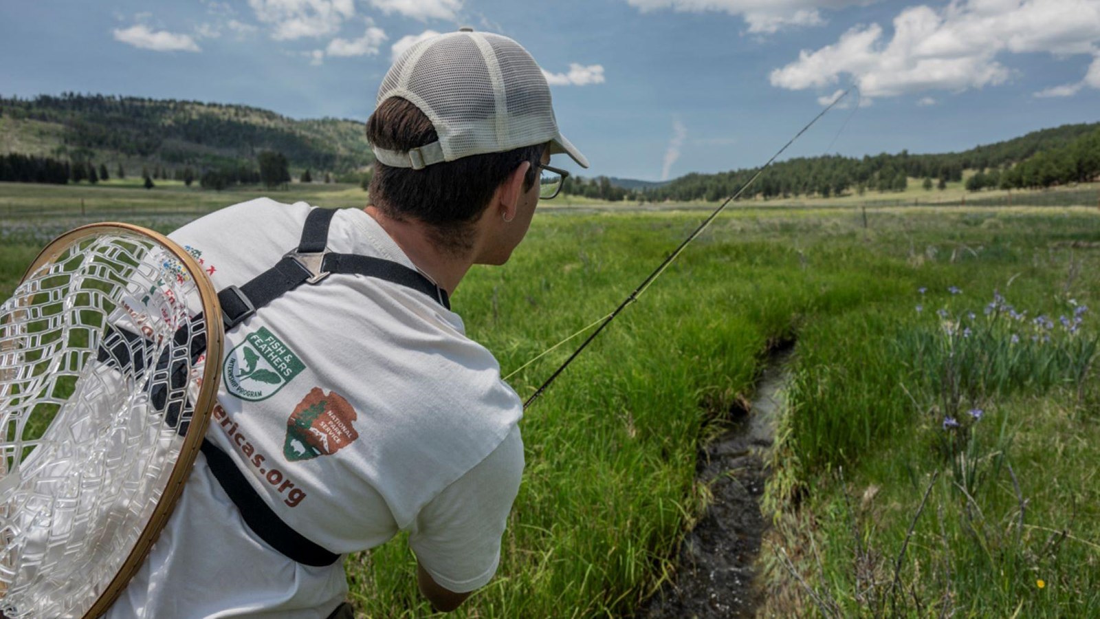 A fisherman holds a flyfishing rod over a very narrow, overgrown creek in a grassland.