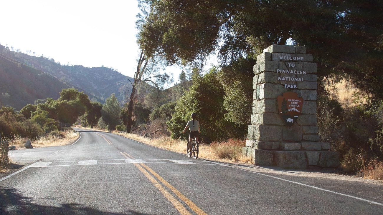Visitor seen along a paved roadway riding a bicycle past a park service sign made into a rock pillar