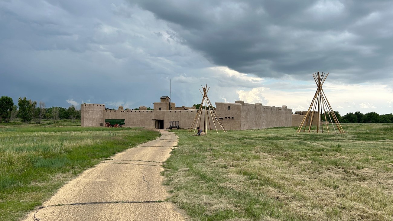 A paved path leads to an adobe fortress. Two tipis are set up in the foreground