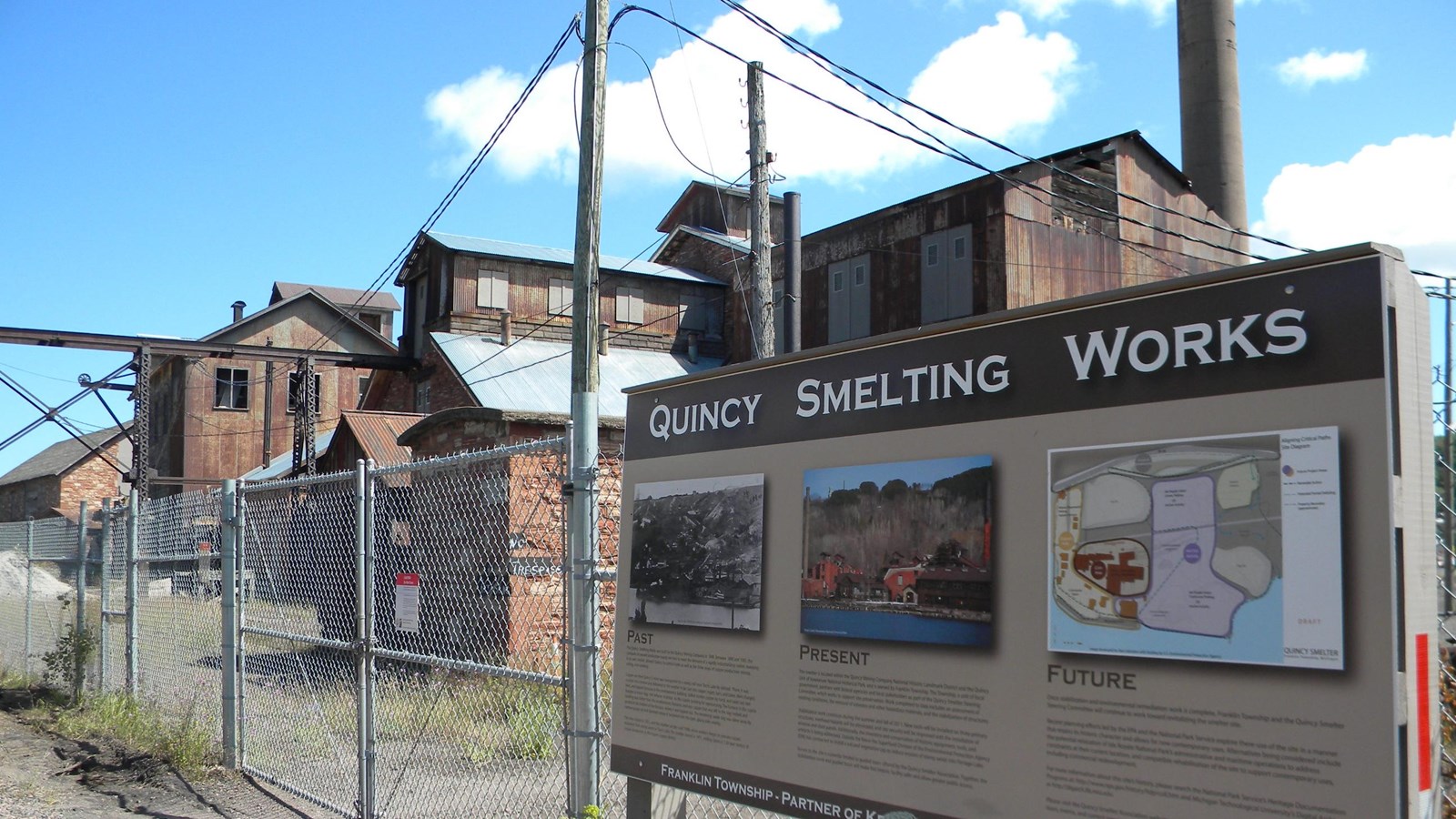 Boarded up industrial buildings made of stone and brick are within a chain link fenced in complex.