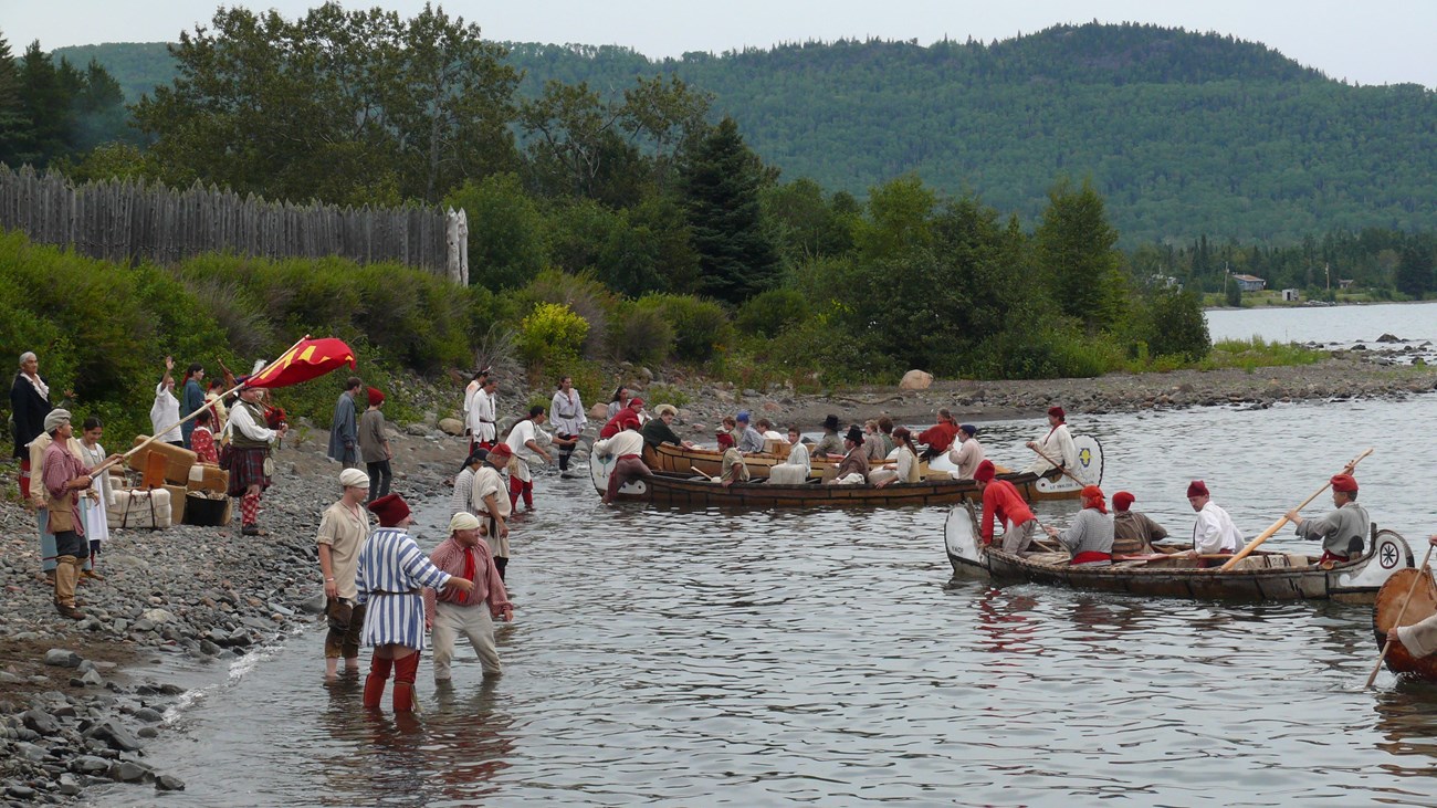 Canoes and people wearing historic clothing at the edge of a lake, next to a stockade.