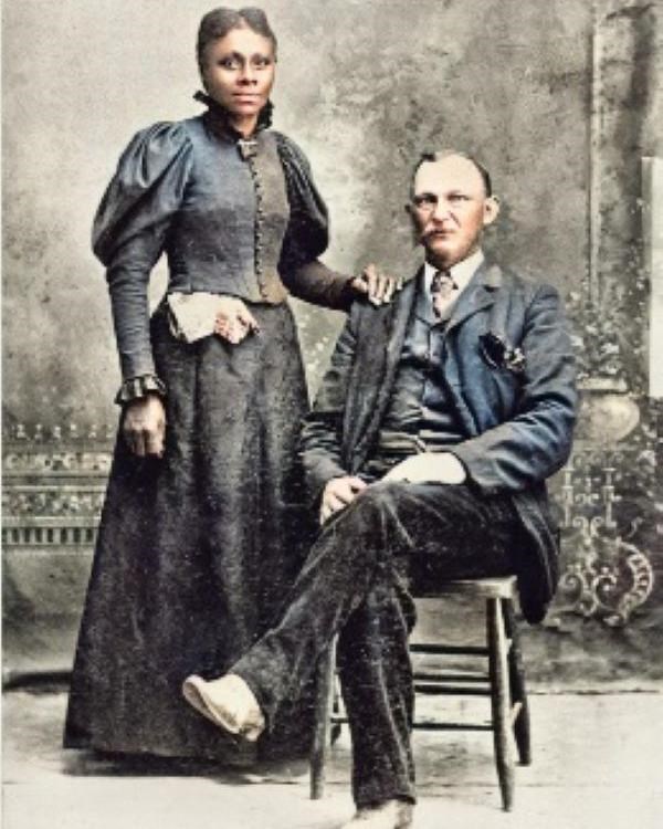 White middle aged man sits wearing suit and tie as black woman in dress rests hand on his shoulder.