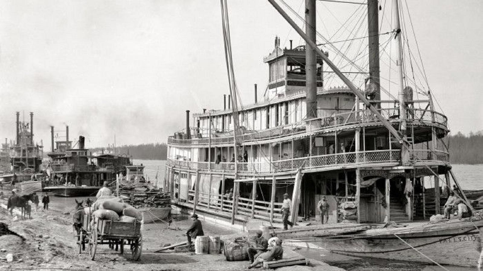 Black and white photo of a steamboat being loaded at a wharf on the river