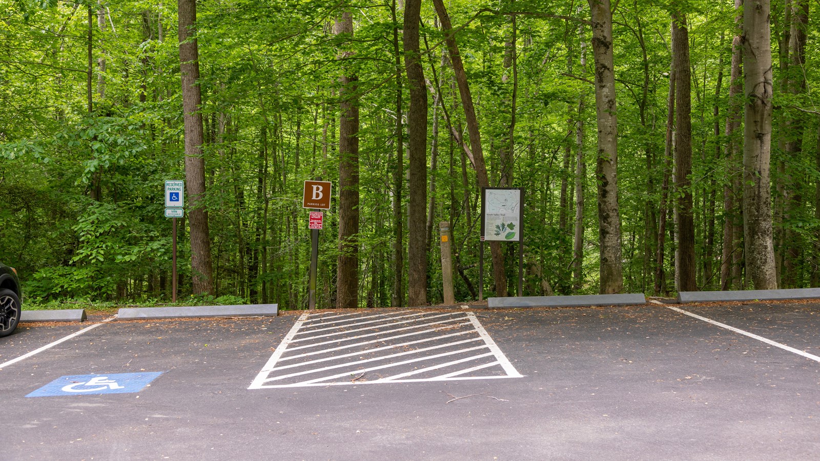 A brown sign with the letter B stands next to a dirt trail and a parking lot