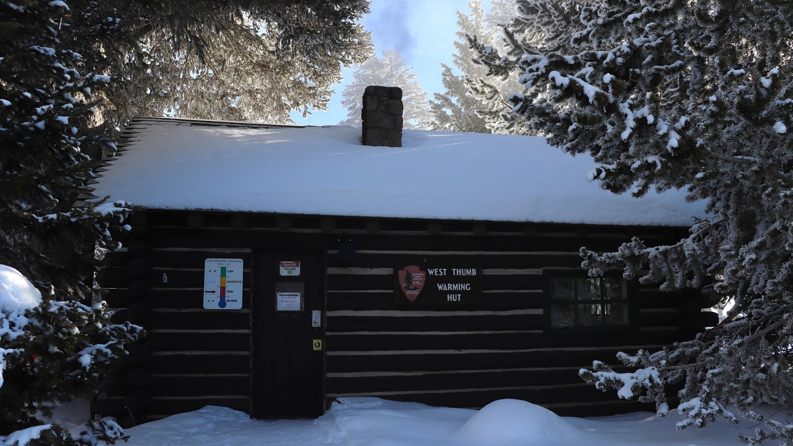 Smoke rises out of the chimney of a snow-covered log cabin building.