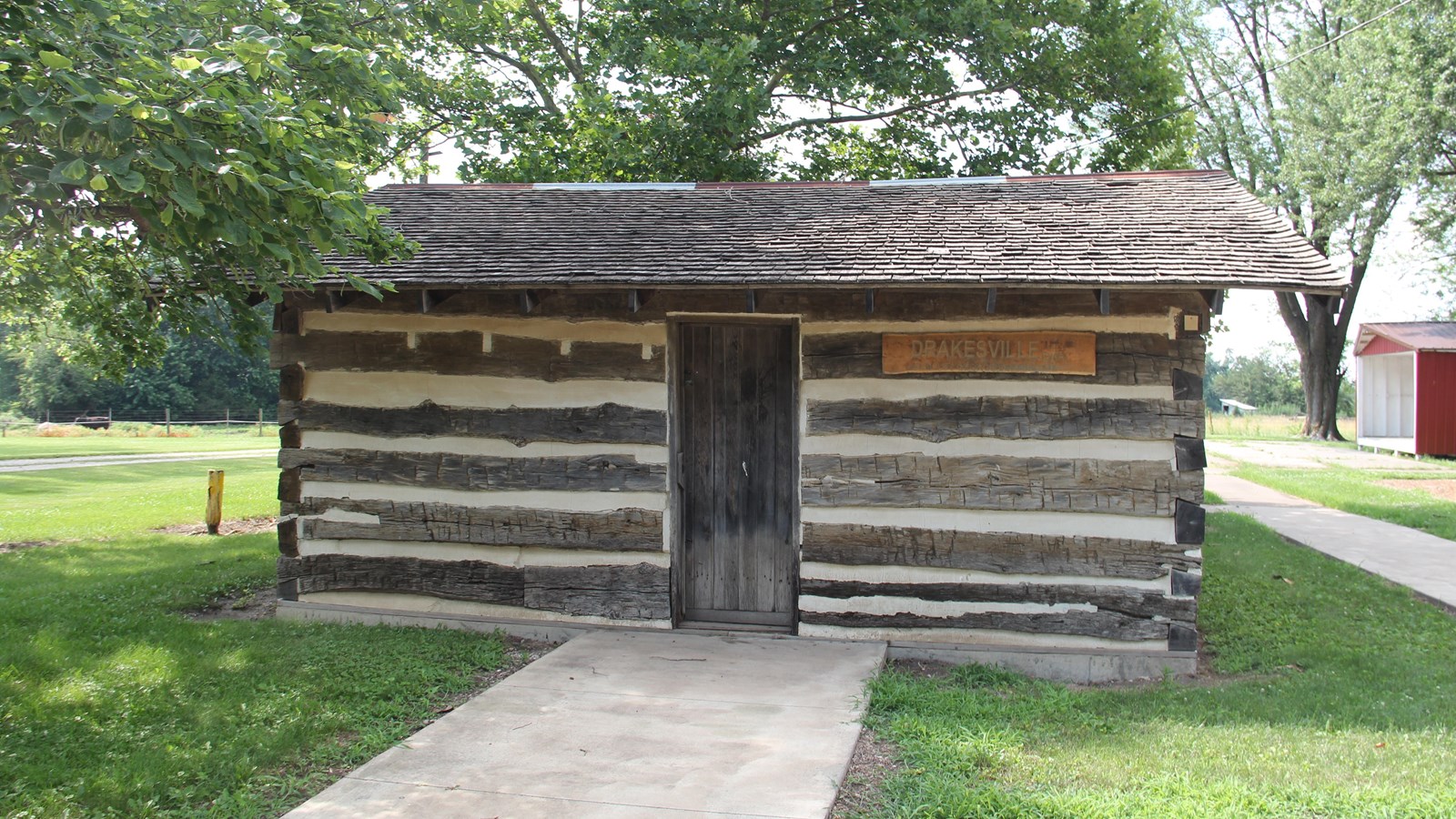 A path leads to the door of a small brown log cabin, surrounded by a grassy city park.