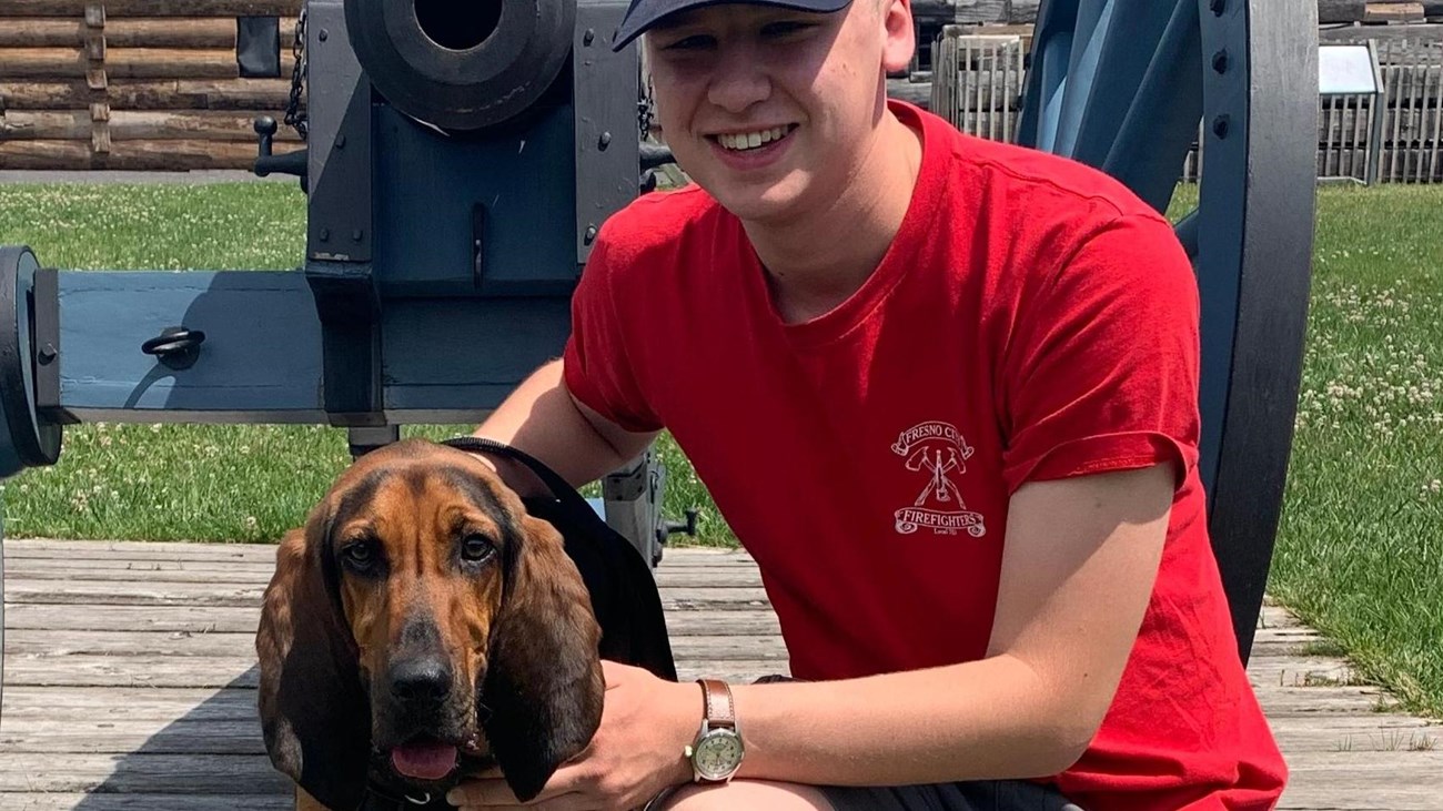A sweet doggy stands on a leash with his person in front of a cannon.