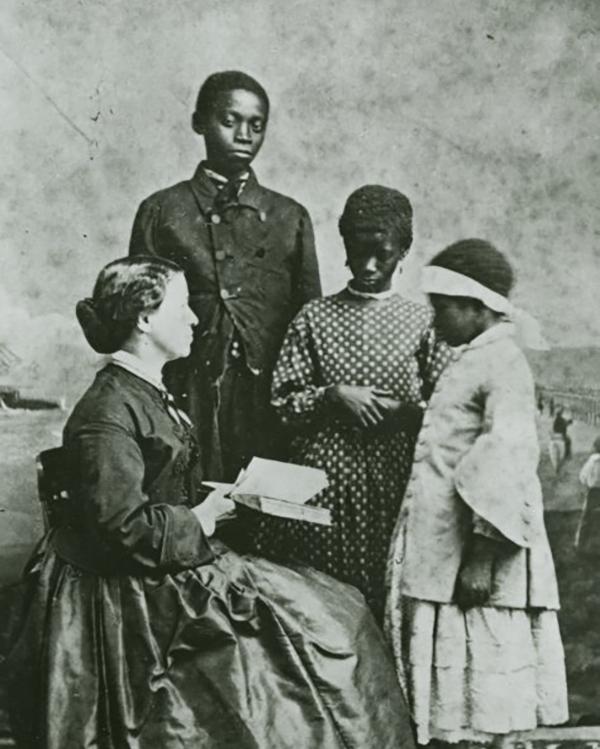 Laura sits with a book while three students stand in front of her.