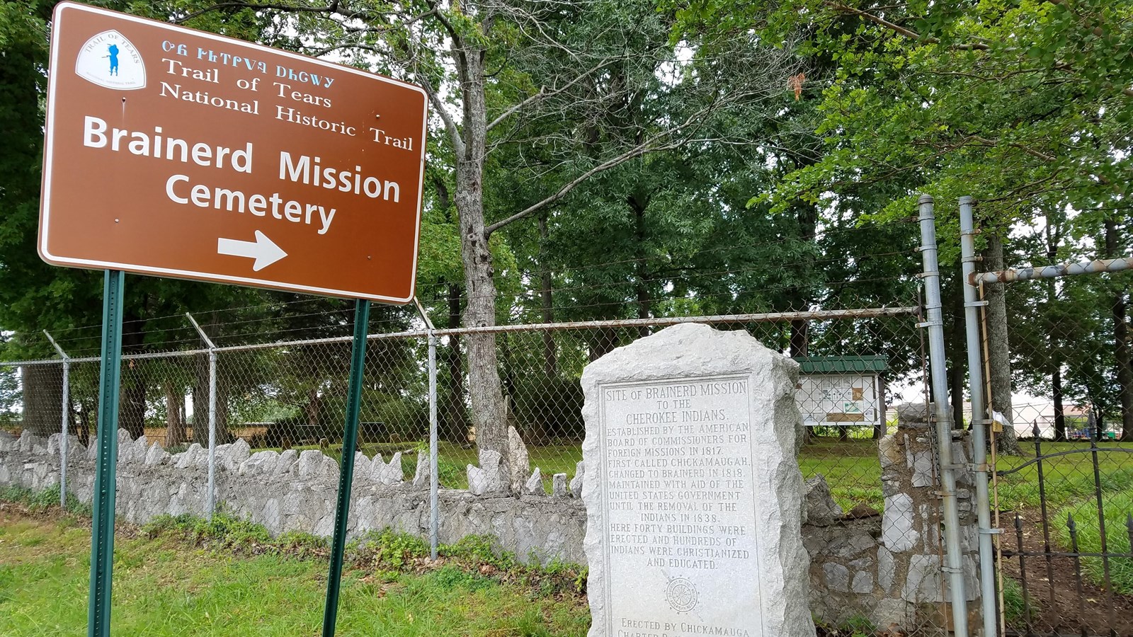 A large brown sign next to a stone marker, in front of a cemetery.
