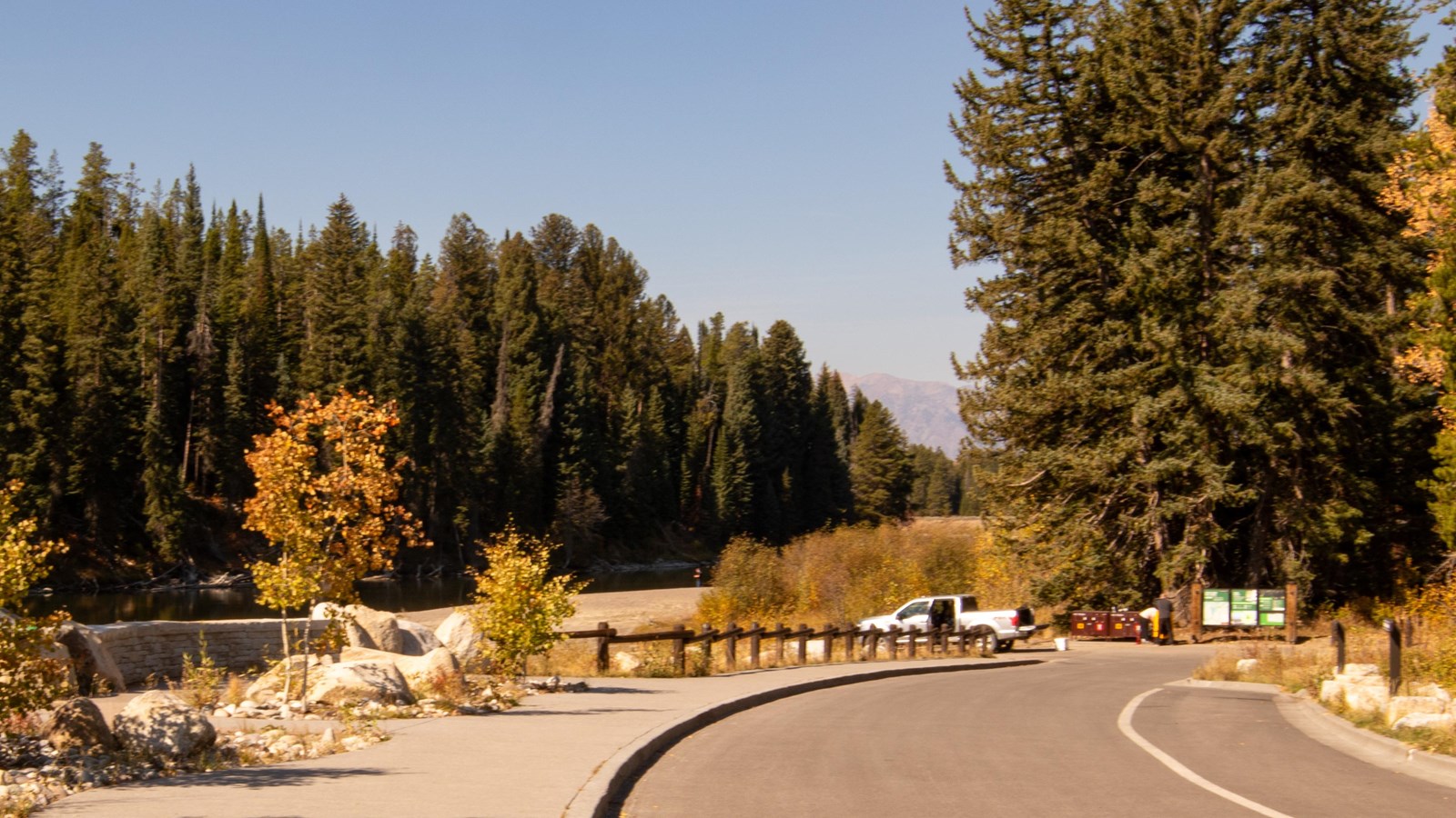 A paved road leads to a boat ramp where a truck unloads. There are trees and river in the back. 