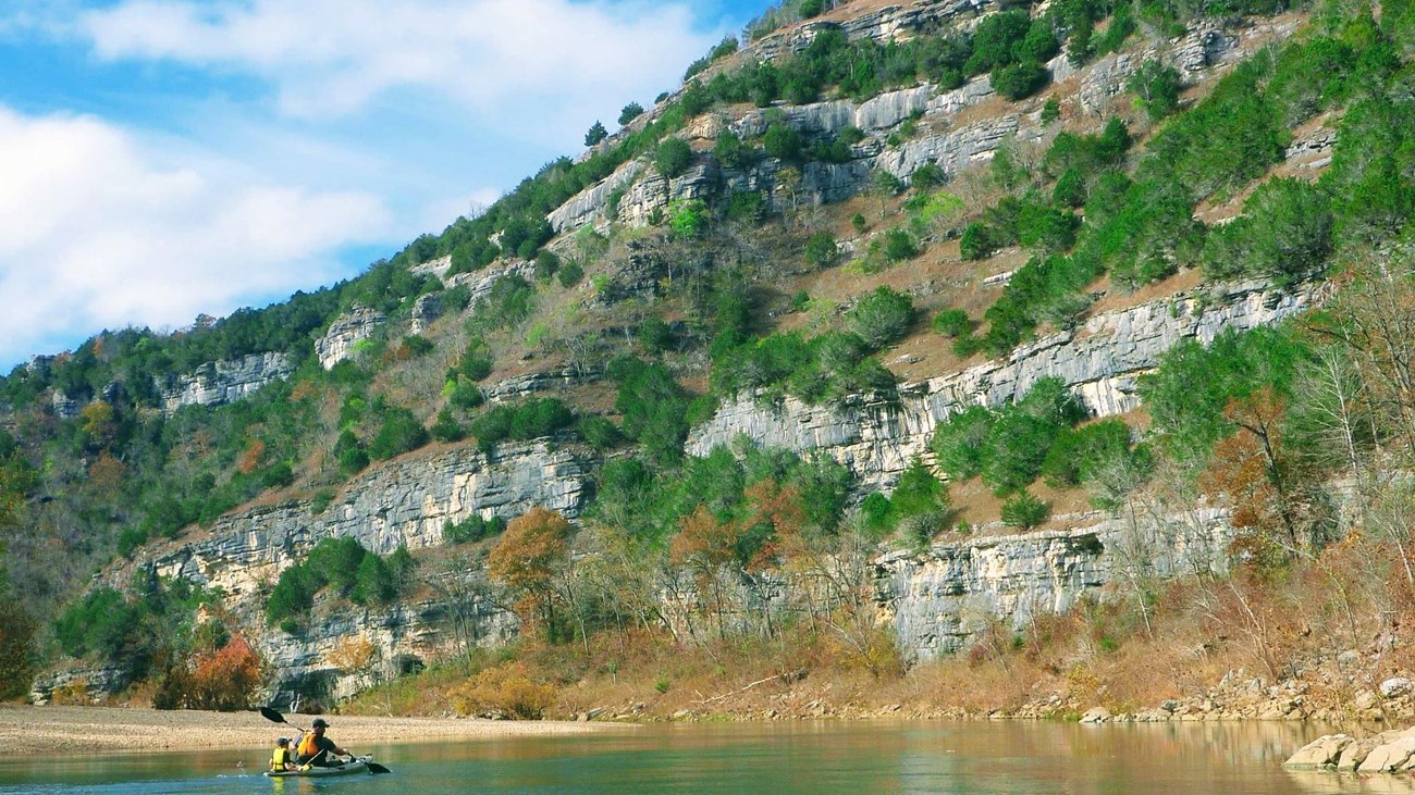 Two paddlers in a kayak passing by a rocky and vegetated bluff.
