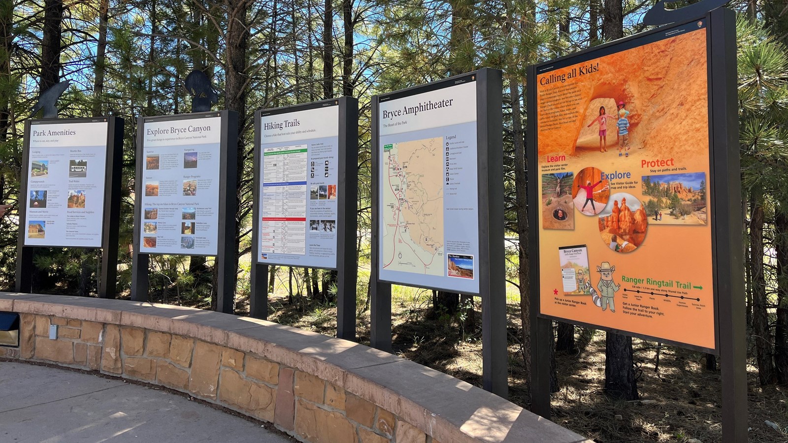 Five informational signs stand side by side behind a curved stone wall.